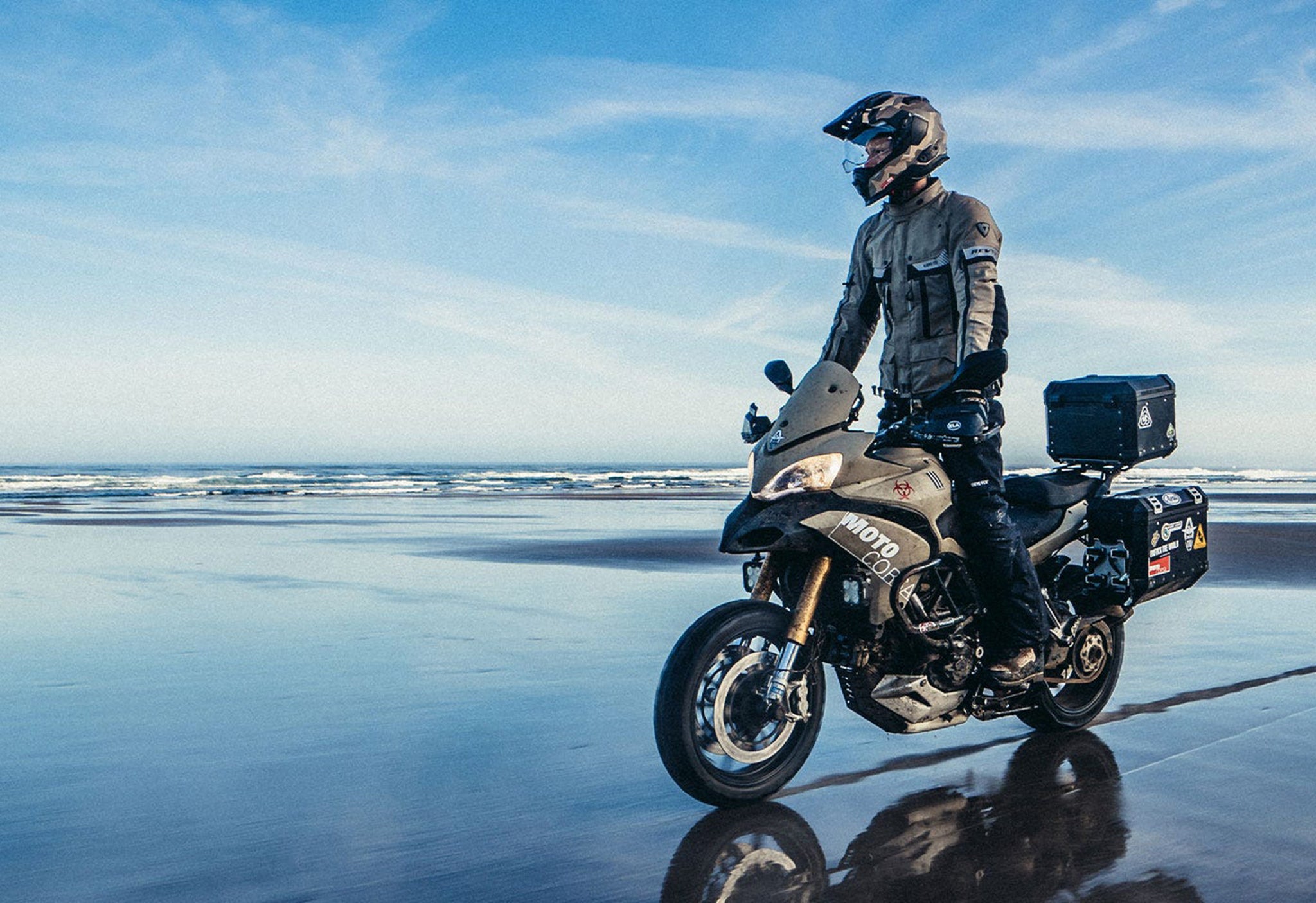 Lifestyle image of a motorcyclist wearing REV'IT! motorcycle gear, while riding an adventure bike on a beach