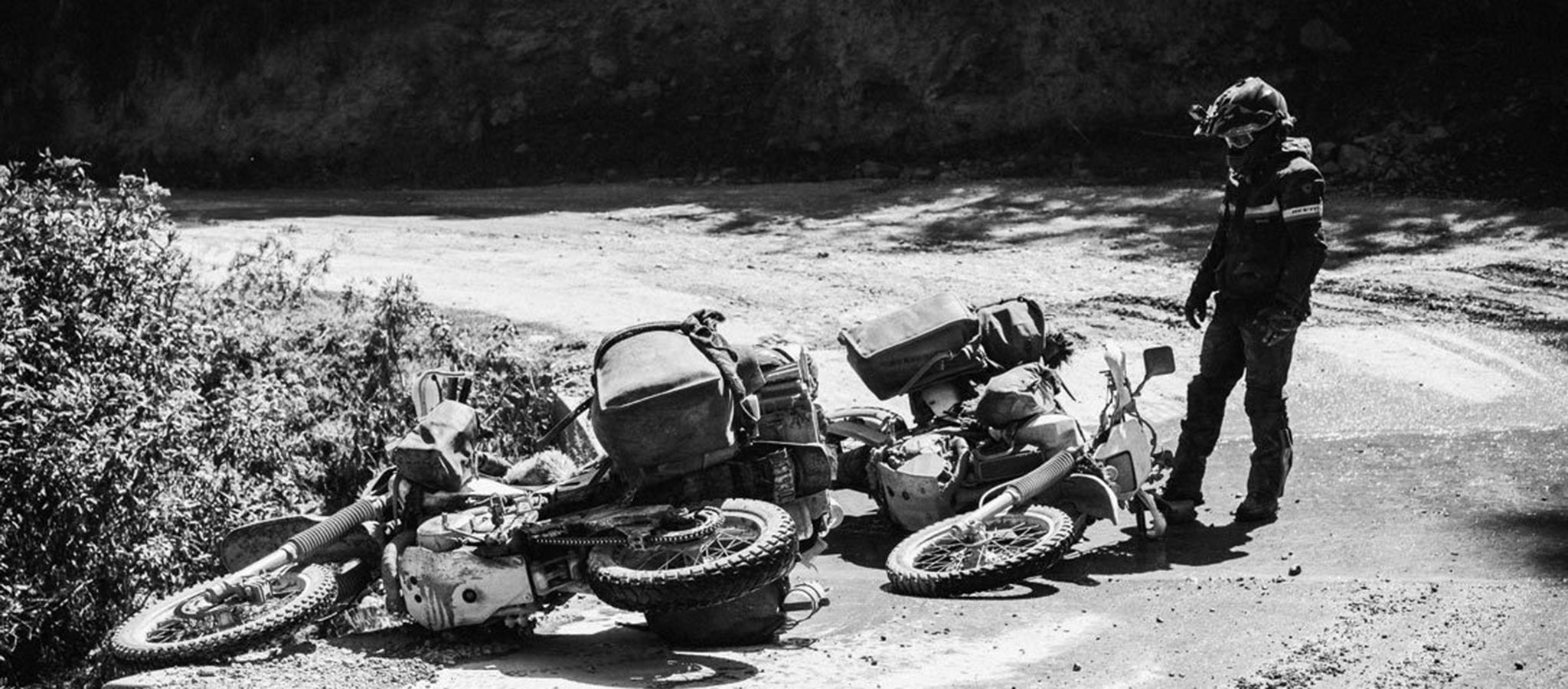 Black and white lifestyle image of a motorcyclist looking at two adventure bikes tipped over on a road