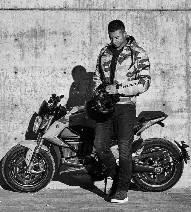 Black and white lifestyle image of a motorcyclist wearing REV'IT! motorcycle apparel, standing next to his motorbike